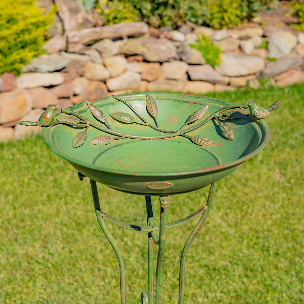 Green birdbath bowl with two bird figures and leafy metal detailing, shown against a garden stone wall.