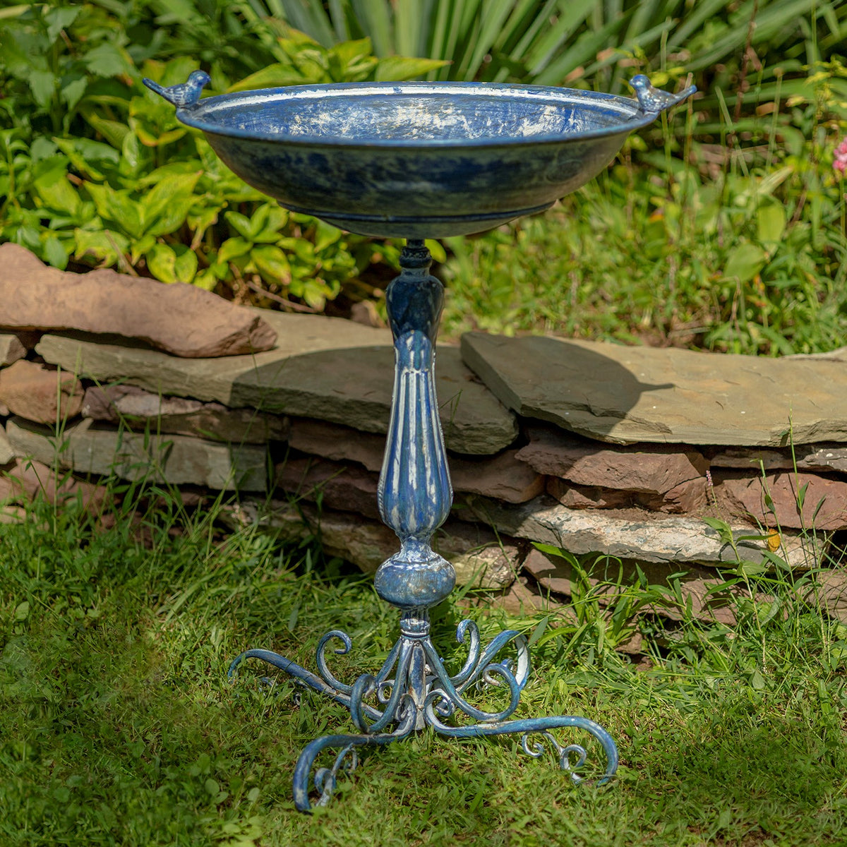 Blue pedestal birdbath with two small bird figures on the rim, shown against greenery and a stone wall.