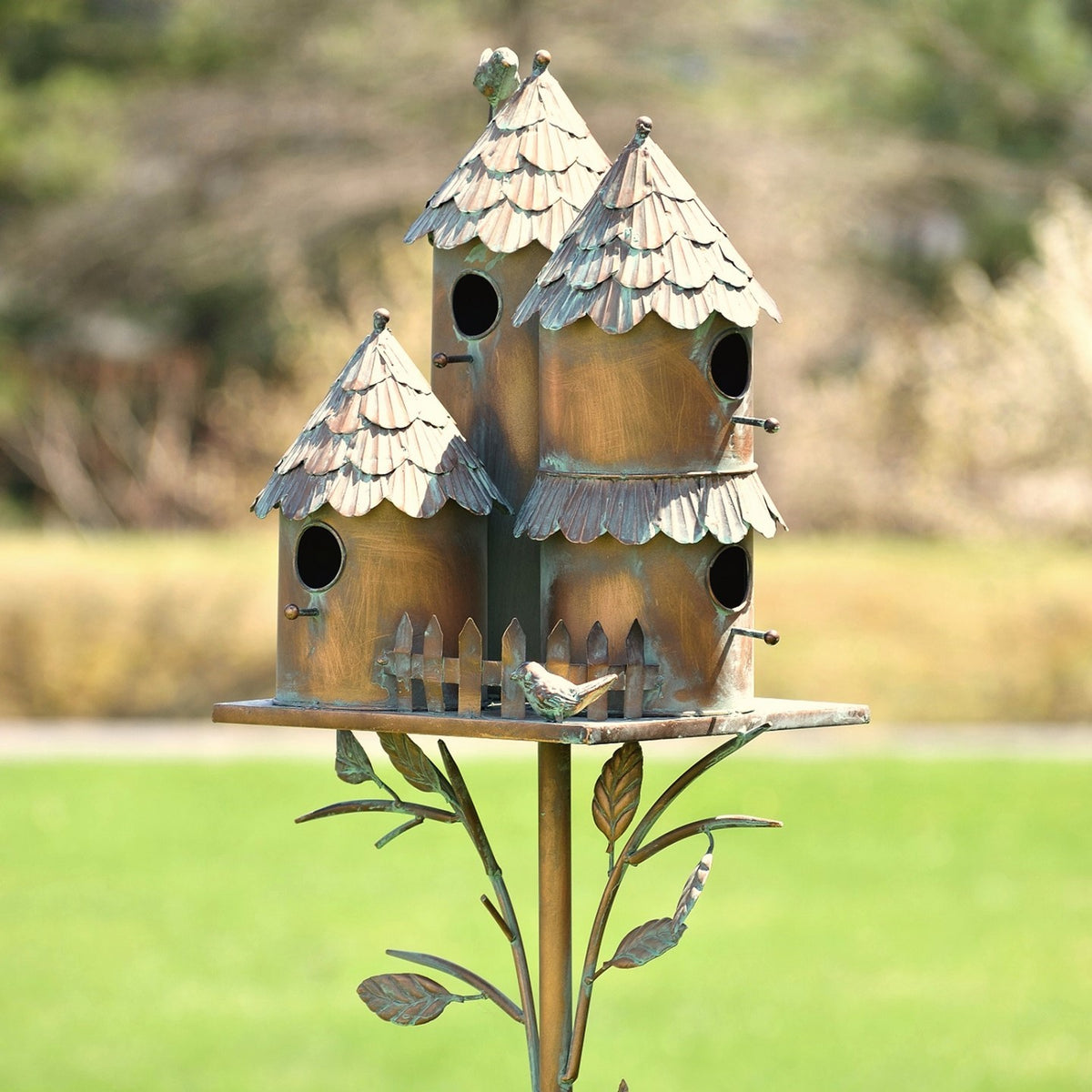 Three rustic metal birdhouses with scalloped roofs sit on a leafy stake in a sunny garden.