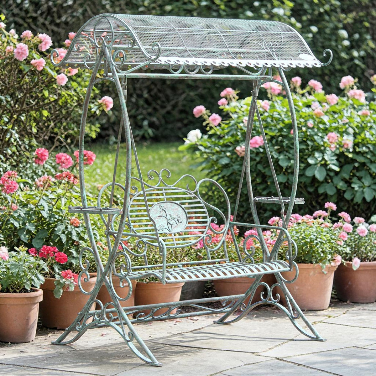 A metal garden swing with a rounded mesh canopy sits on a paved patio among flowering pots.