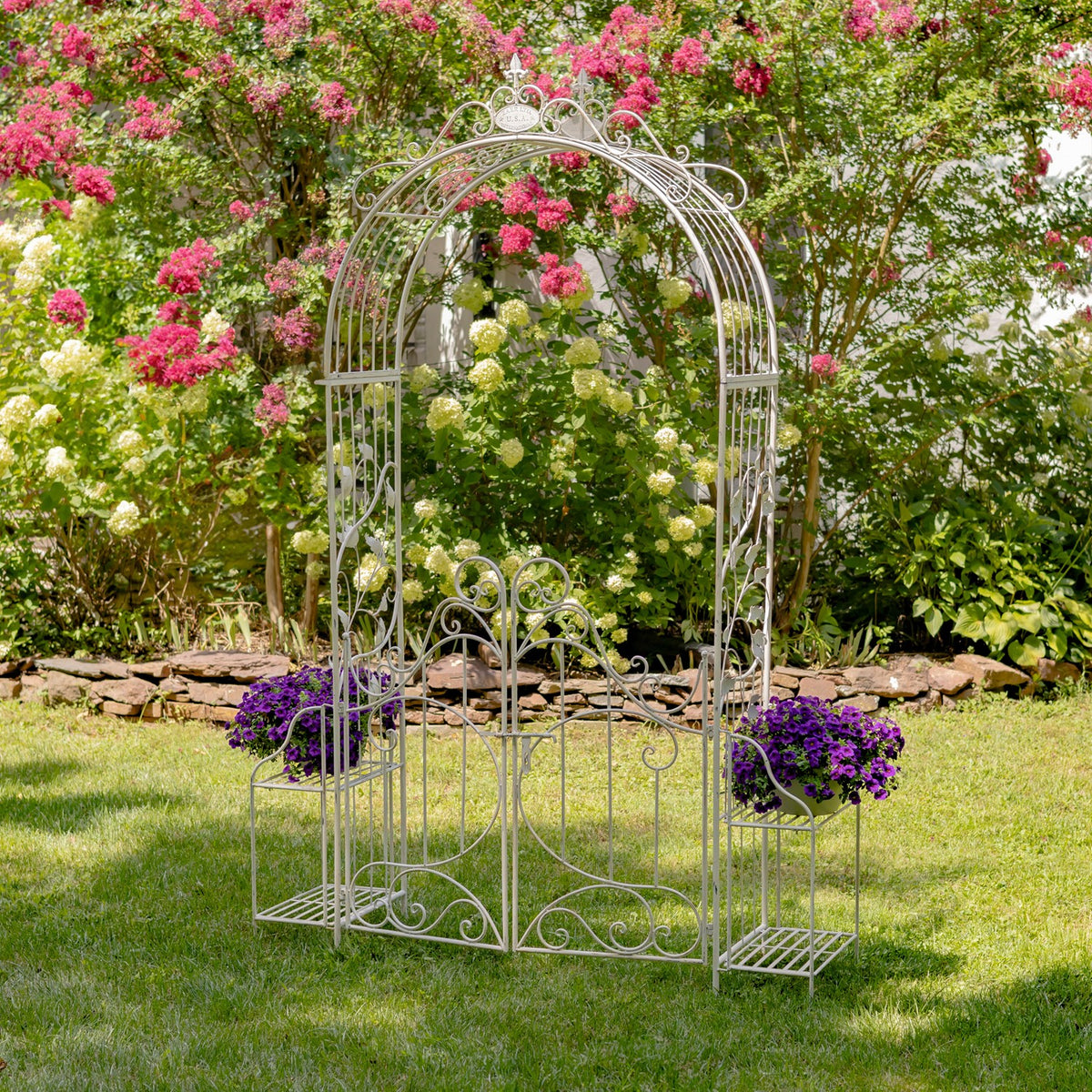 Antique white iron garden gate arch with side plant stands among flowering shrubs