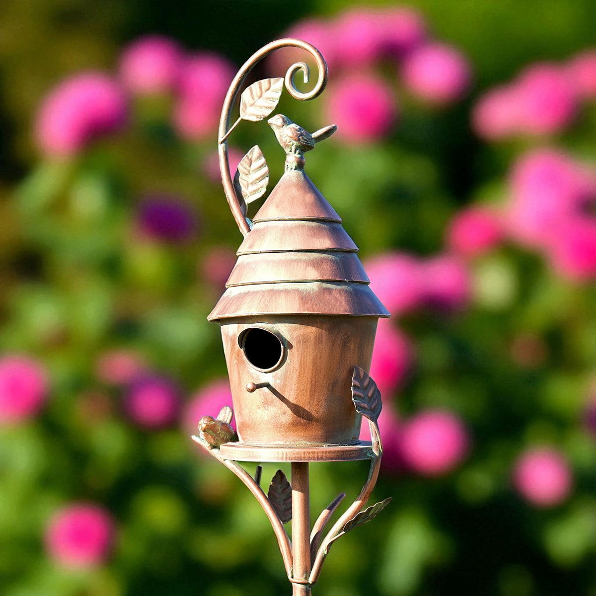 Copper birdhouse stake with a tiered roof and small bird ornaments, set against blurred pink flowers.