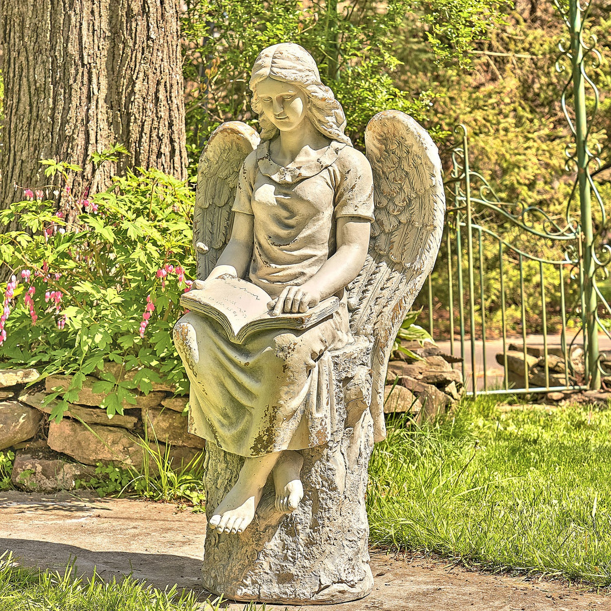 A seated angel statue with wings reads an open book beside a garden path.