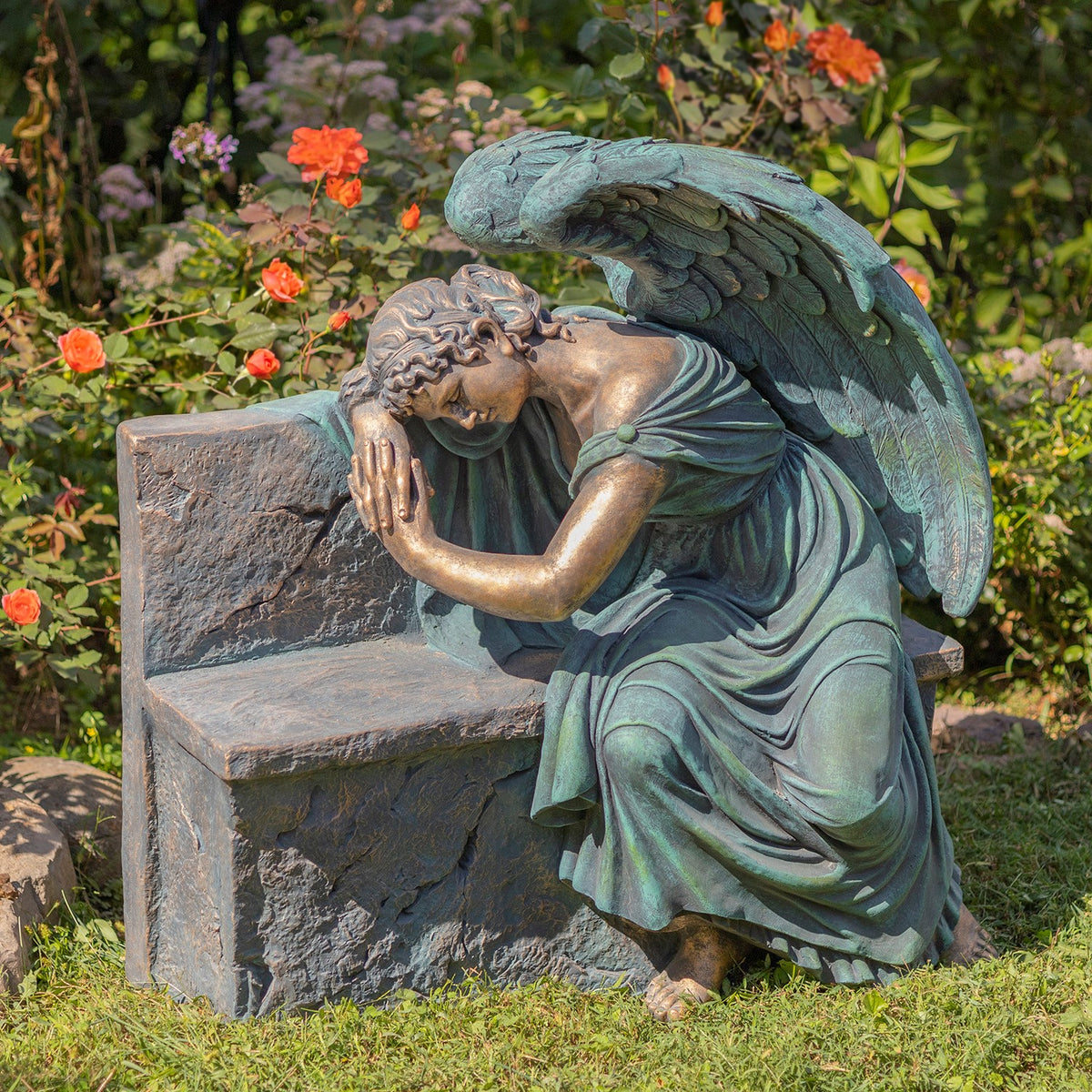 Bronze-toned angel rests against a stone bench amid flowering garden plants.