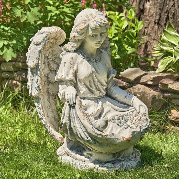 Seated angel statue with birds in its lap, shown among garden plants and stones.