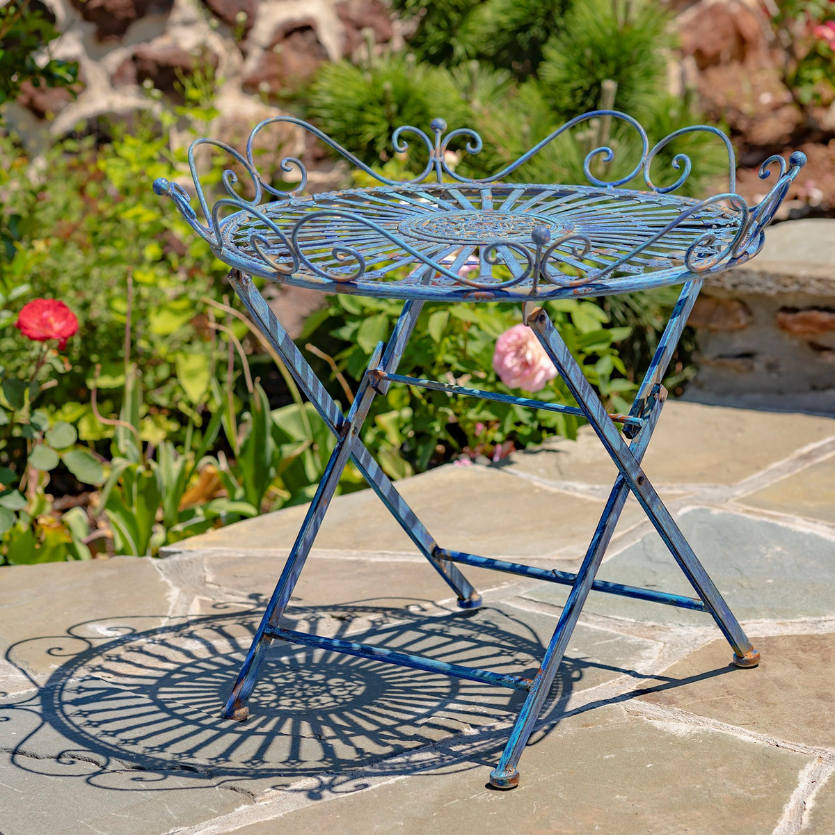 Blue folding iron table with a circular scrollwork top, photographed on a stone patio.