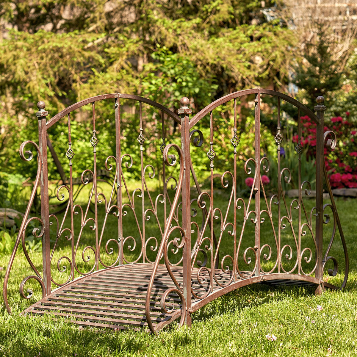 Ornate arched metal garden bridge with a slatted walkway on a lawn among shrubs and flowers.