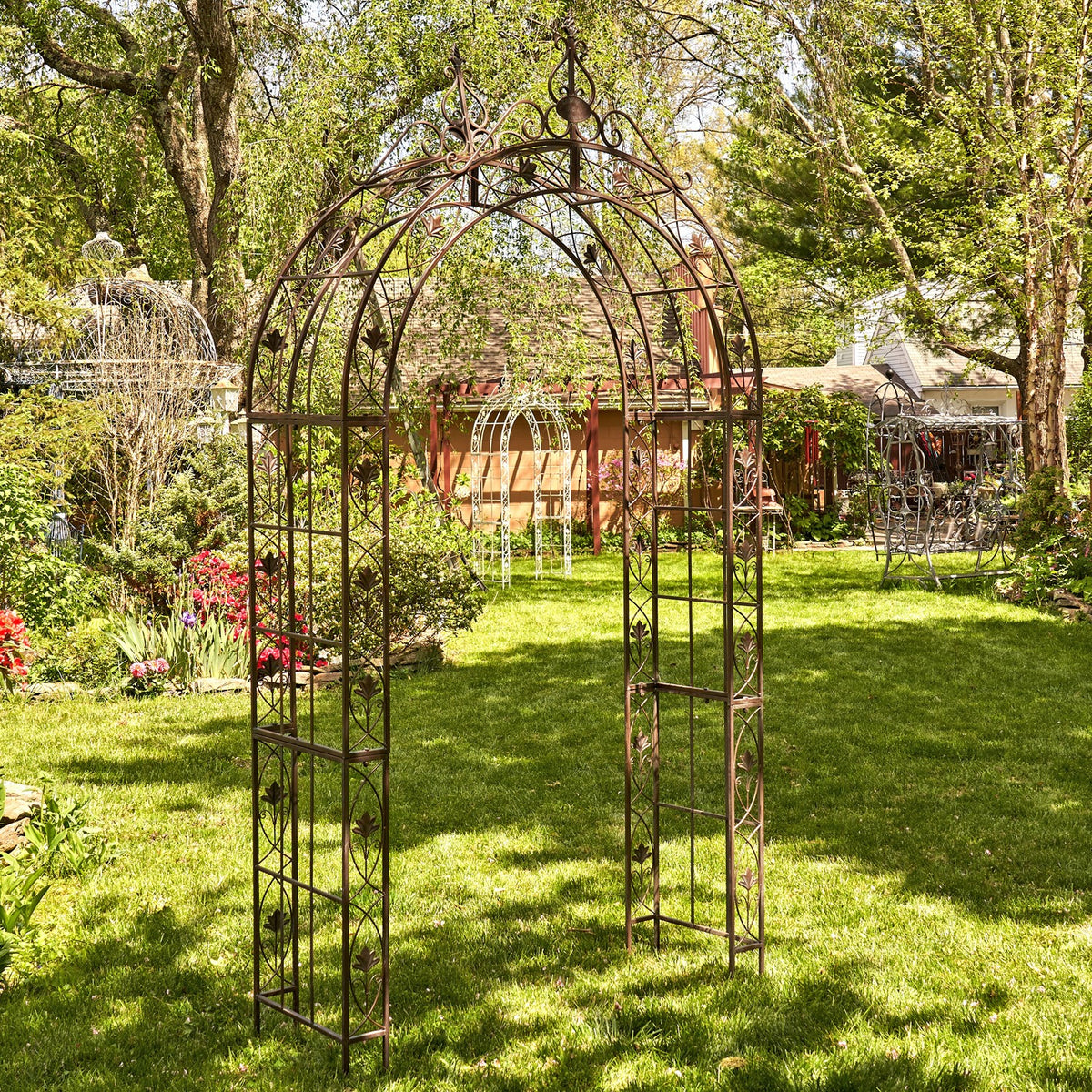 Dark metal garden arch framed by trees and flowers, with another arch visible behind it.
