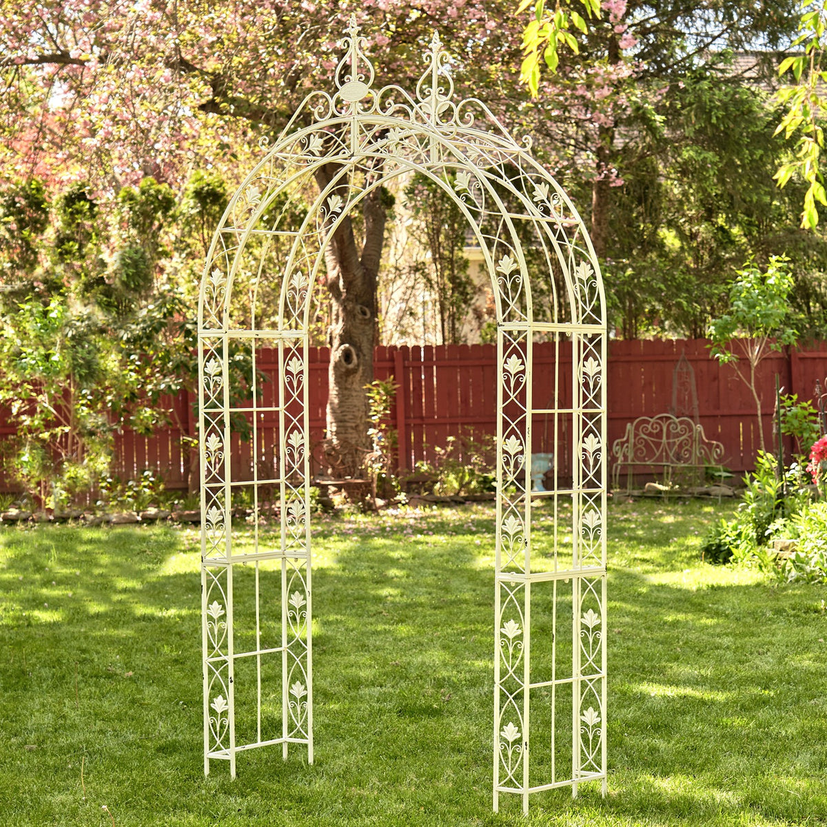 Ornate pale metal garden arch standing on a lawn beneath flowering trees.
