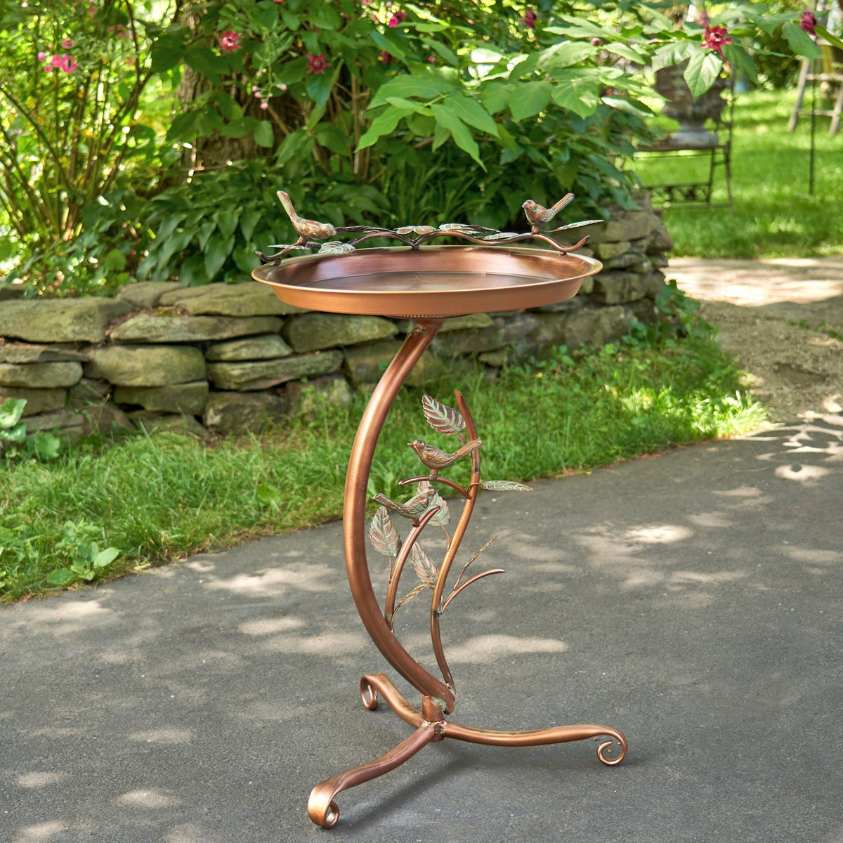 Copper-toned birdbath with bird and leaf ornaments on a curved branch-style stand, beside a stone garden wall.