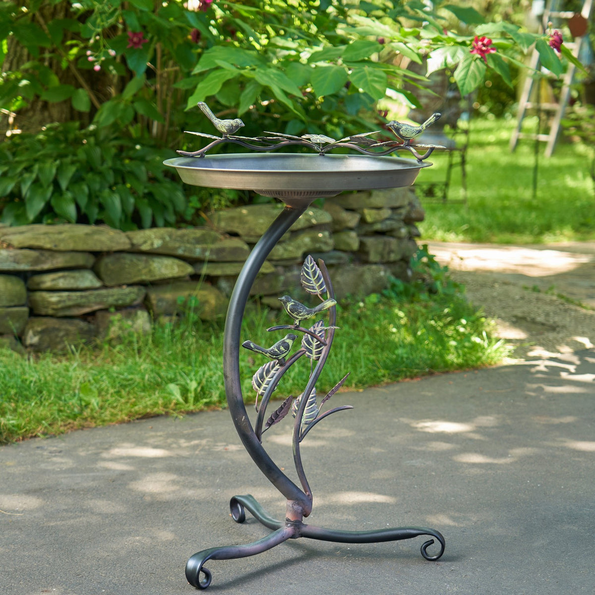 A shallow metal birdbath on a curved branch-style stand with decorative birds and leaves, set beside a garden path.
