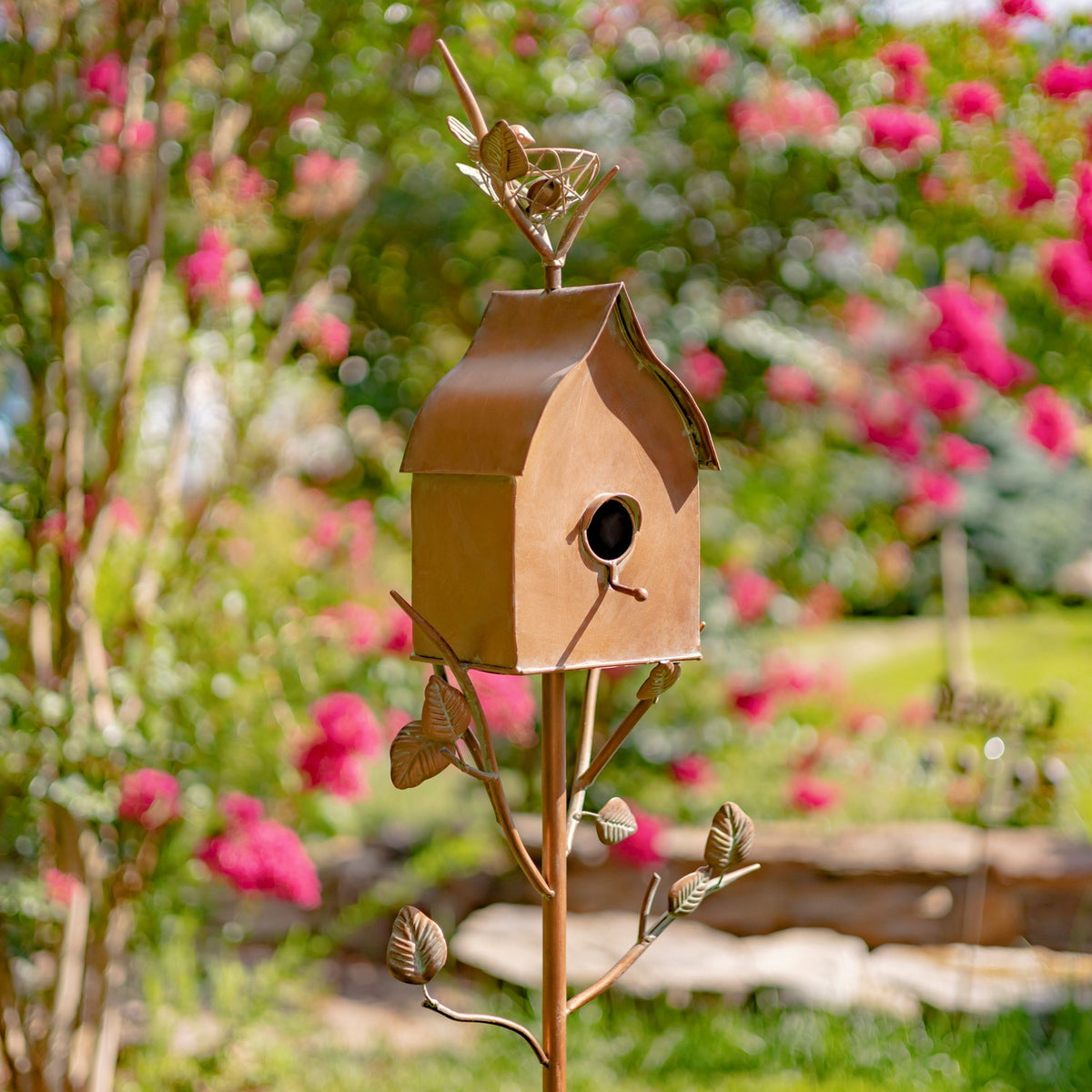 Copper-toned birdhouse stake with leaf details and a bird ornament stands among colorful garden flowers.