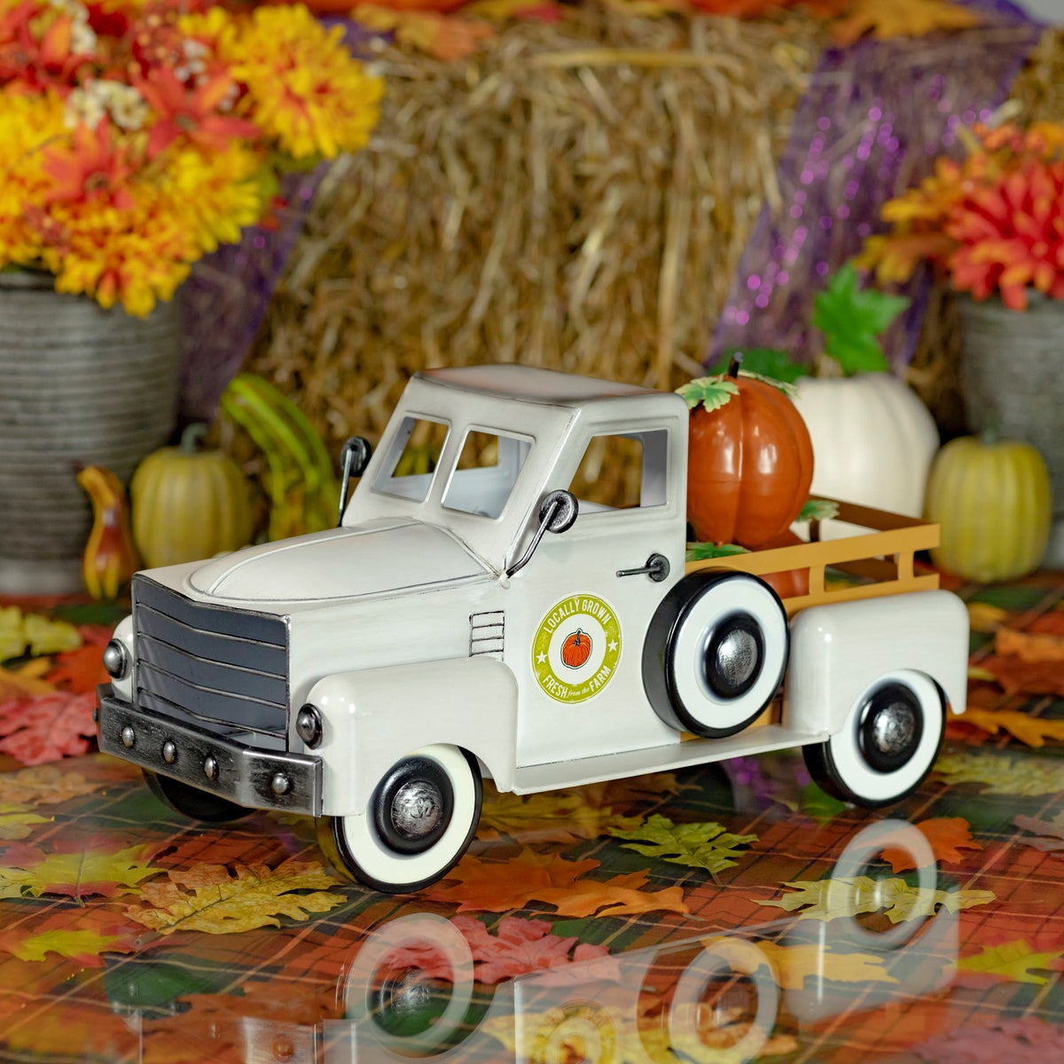 Antique-white pickup truck carrying pumpkins on a leaf-covered tabletop amid autumn decorations.