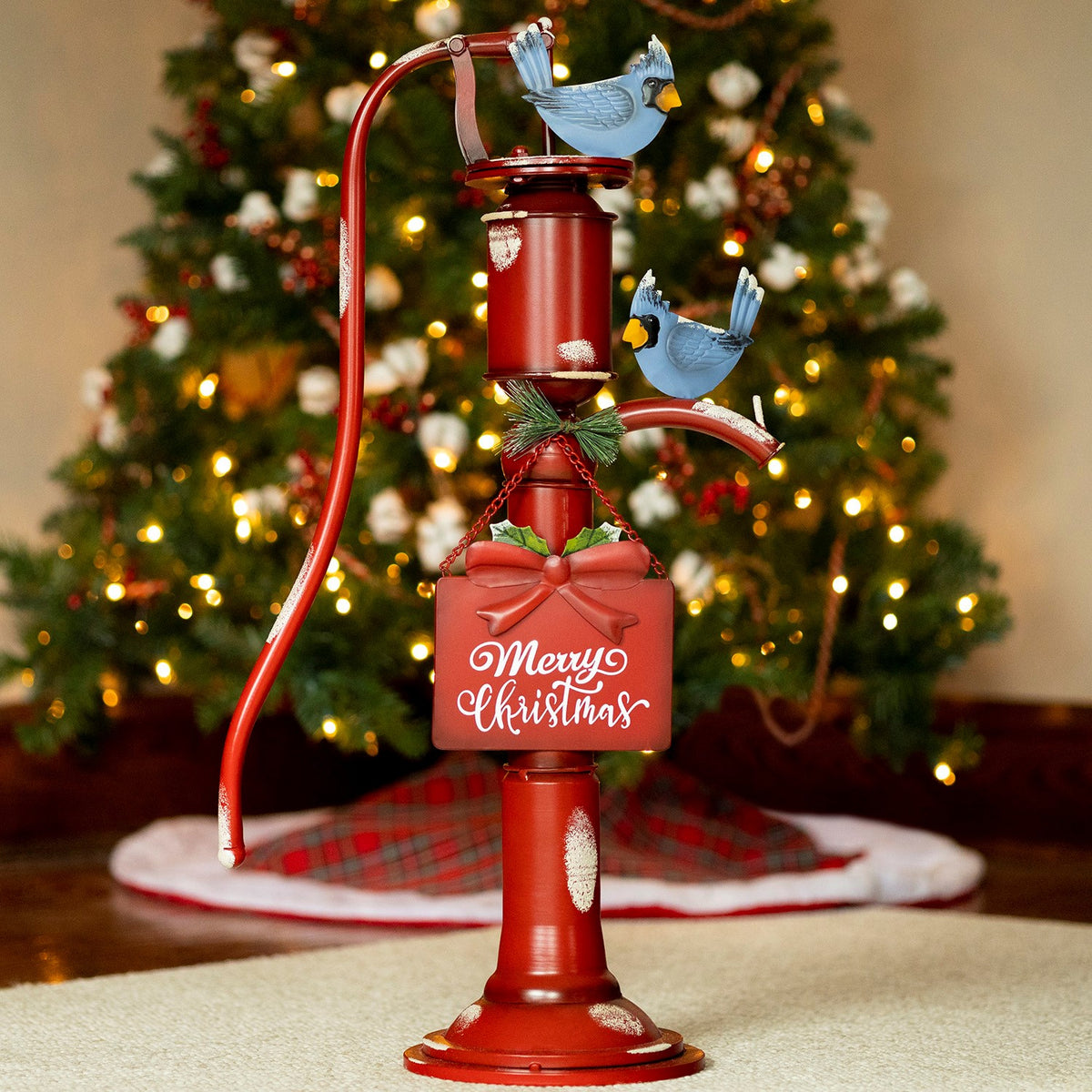 Red vintage-style water pump decor with two blue birds and a hanging sign, displayed before a lit Christmas tree.