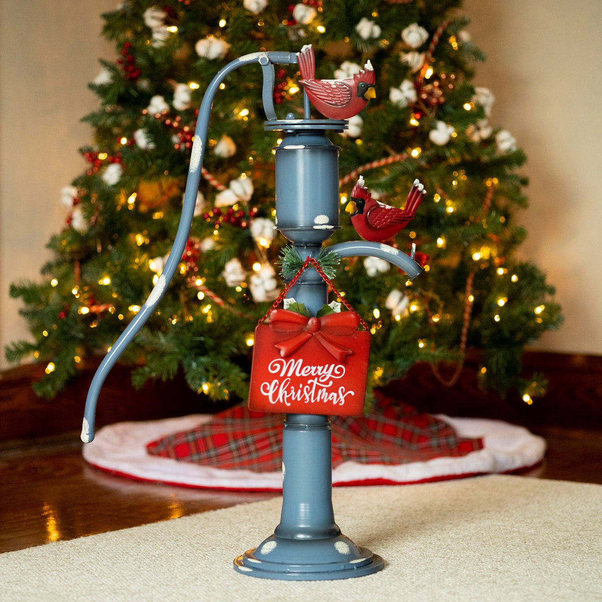 Blue vintage-style water pump with two red cardinals and a hanging holiday sign, in front of a lit Christmas tree.