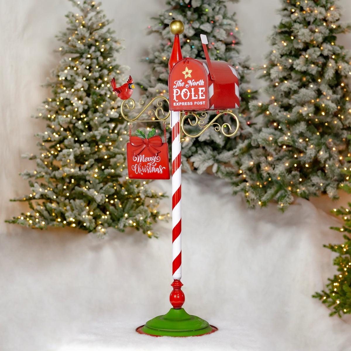Red mailbox on a striped post with a hanging Christmas sign and cardinal, displayed among lit trees.