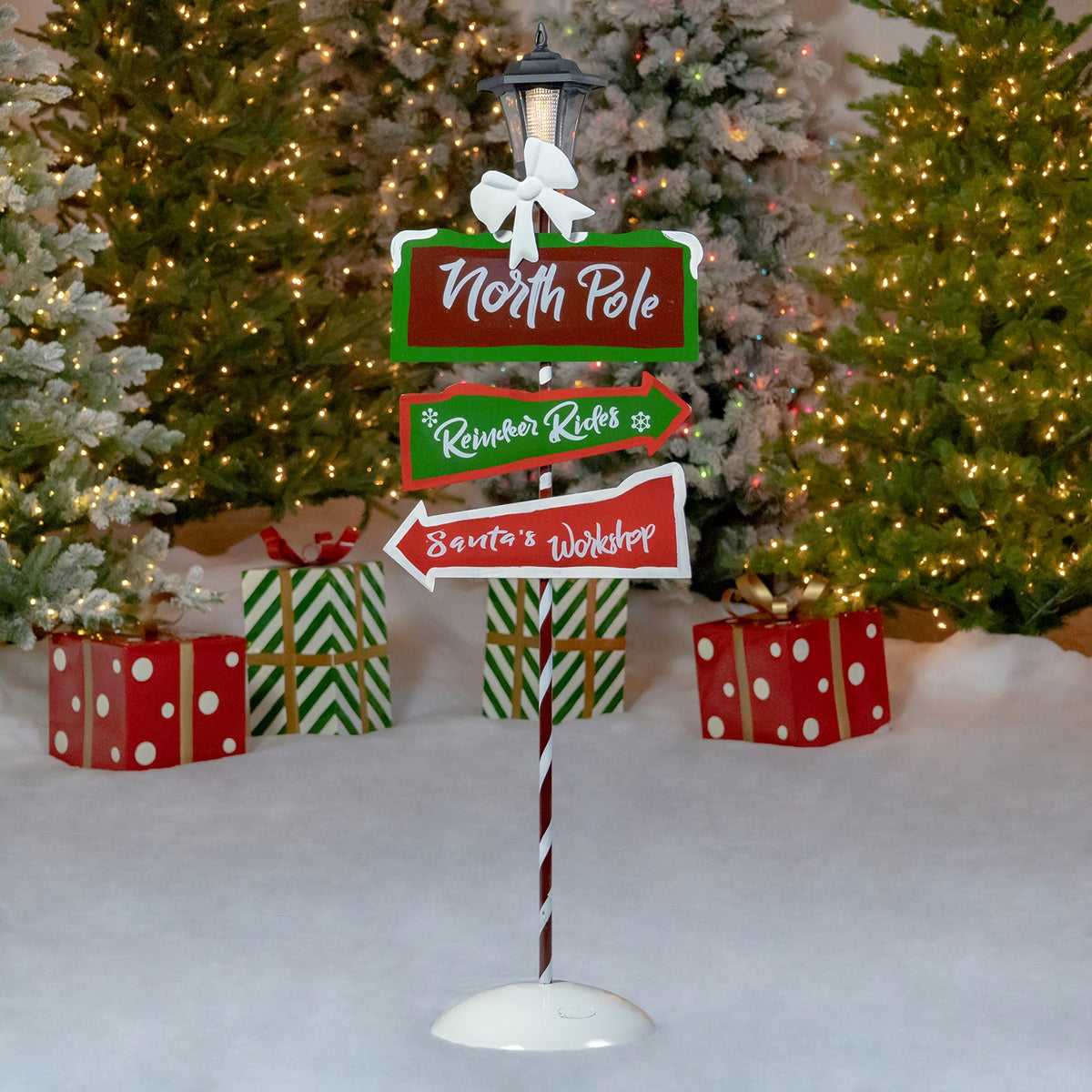 A full candy-cane-striped holiday signpost with a lantern stands in snow among lit trees and wrapped gifts.