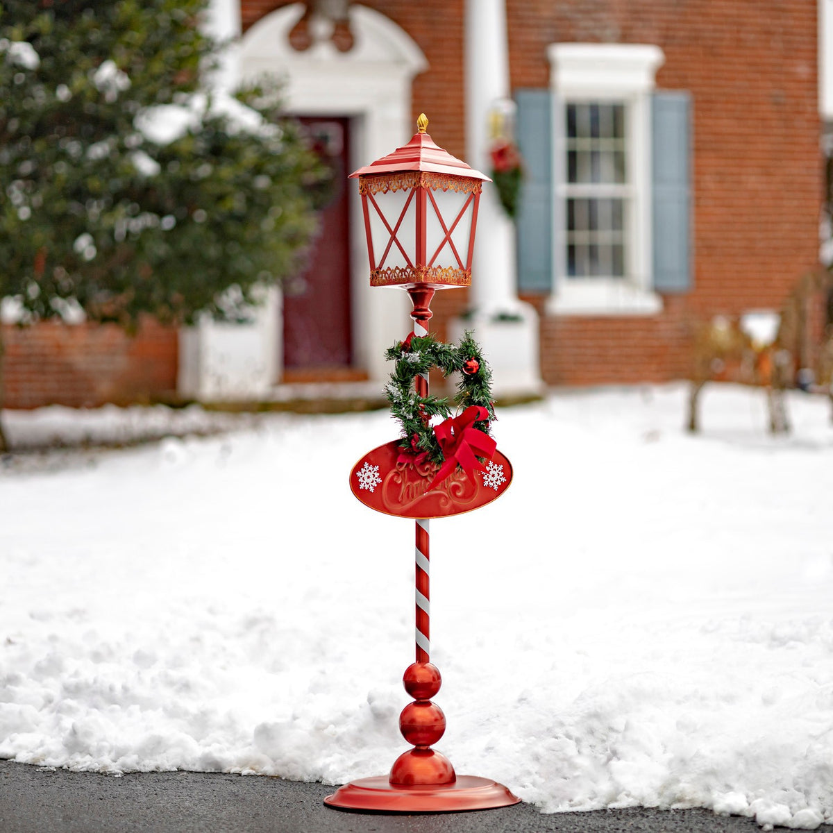 Red standing lantern with a wreath and striped pole set in snow outside a brick house.
