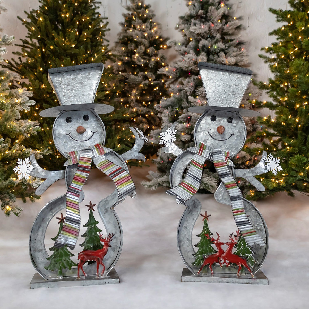 Two galvanized snowman figures with striped scarves, Christmas trees and red reindeer, posed before lit trees.
