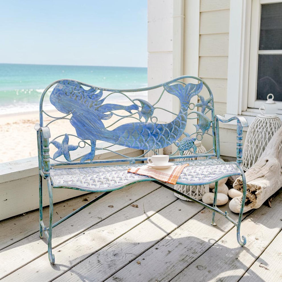 Blue metal mermaid bench on a seaside porch, with a cup and magazine on its curved seat.