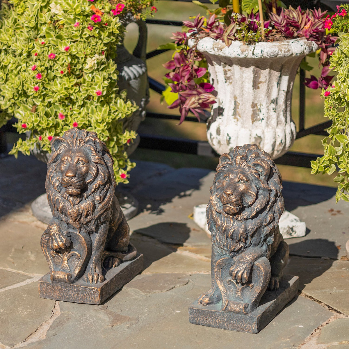 Two seated lion statues flank a flower planter on a sunlit stone patio.