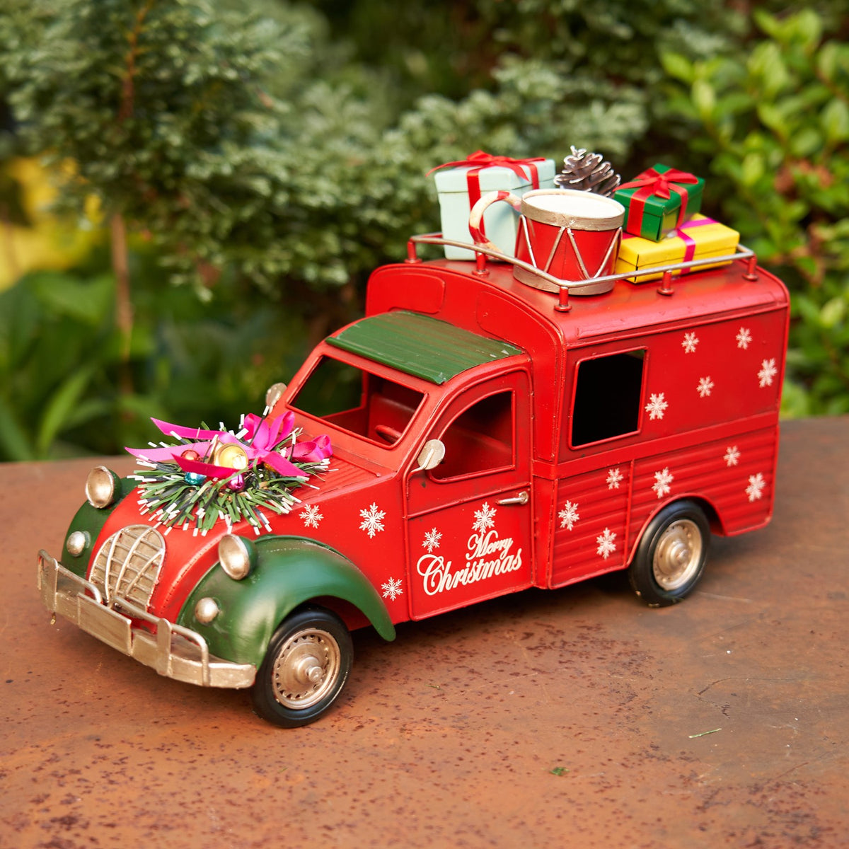 Red vintage-style Christmas truck with snowflakes, gifts and a drum on its roof, shown outdoors.