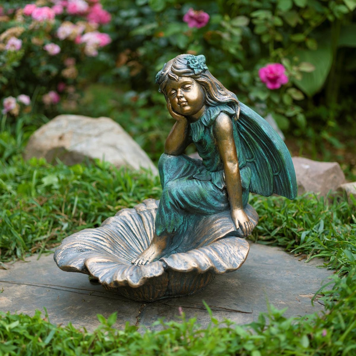 A seated fairy statue rests on a large flower-shaped base among garden stones and greenery.