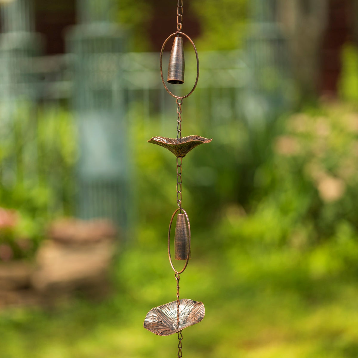 Antique bronze rain chain with mushroom-shaped cups and chimes hanging against a leafy garden backdrop.