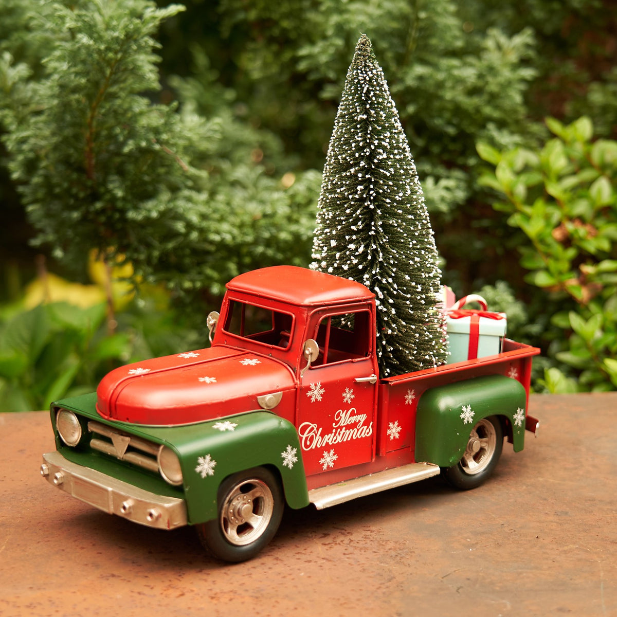 Red and green Christmas truck with snowflakes, carrying a miniature tree in a garden setting