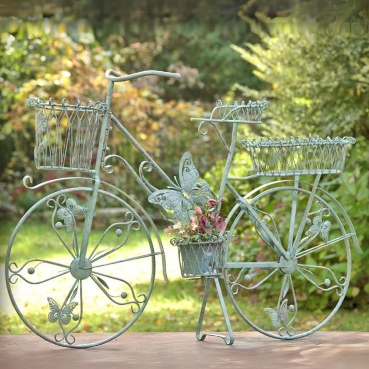 Ornate green bicycle plant stand with flower baskets and pink flowers, displayed outdoors against a garden backdrop.