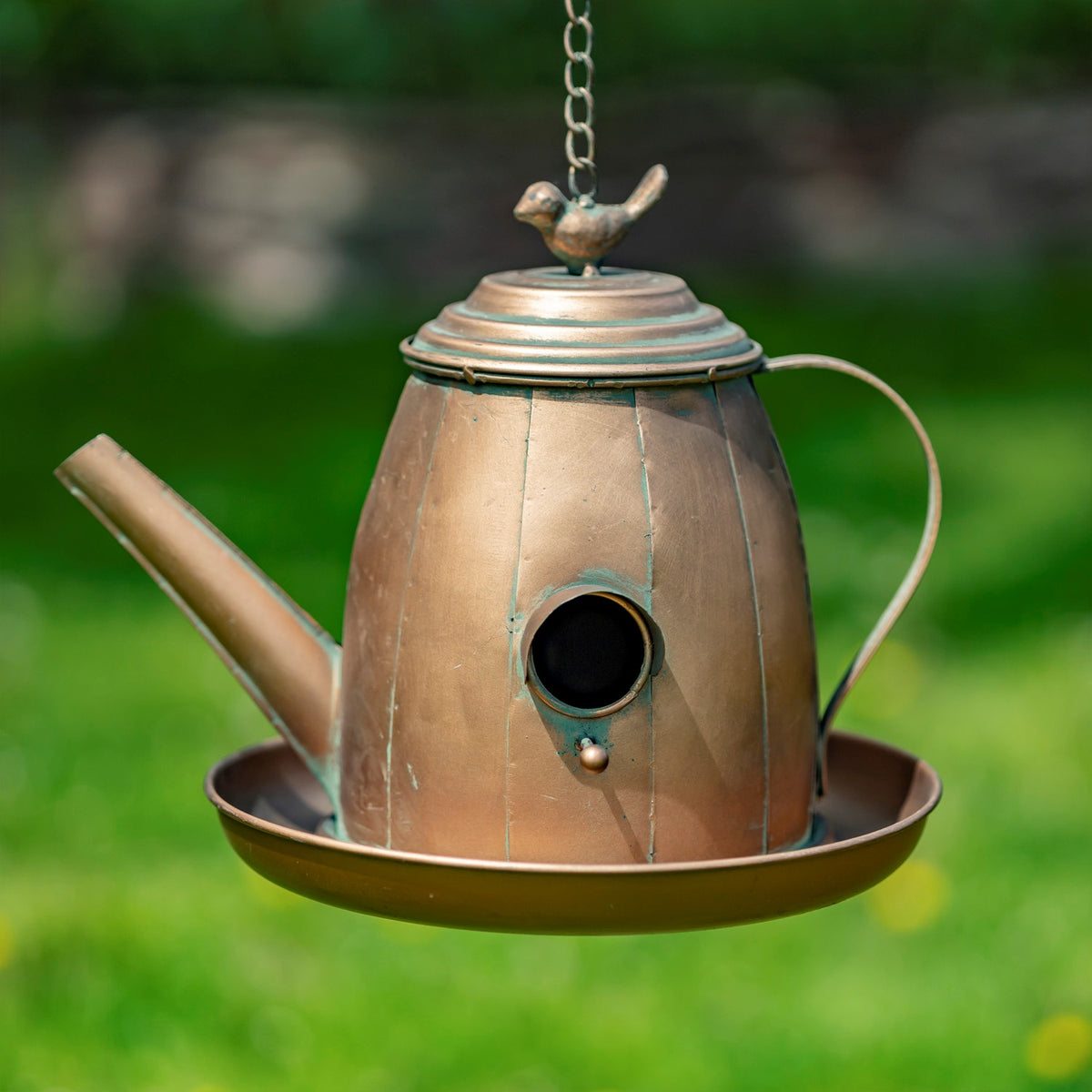 Antique copper teapot-shaped birdhouse with a round entrance hole, hanging chain, and small bird ornament on its lid.