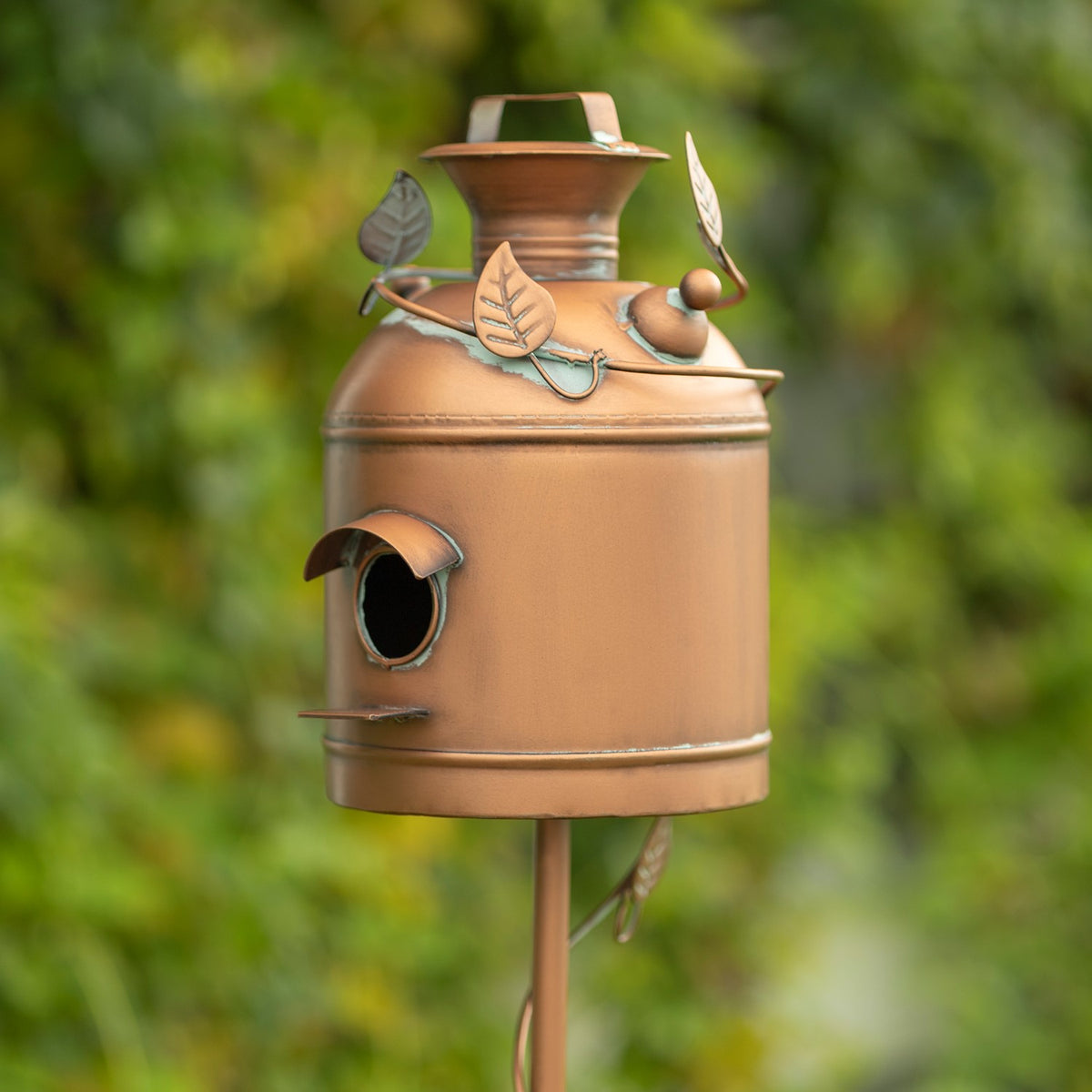 Copper-toned birdhouse with a round entrance, decorative leaves and a milk-can-shaped top.