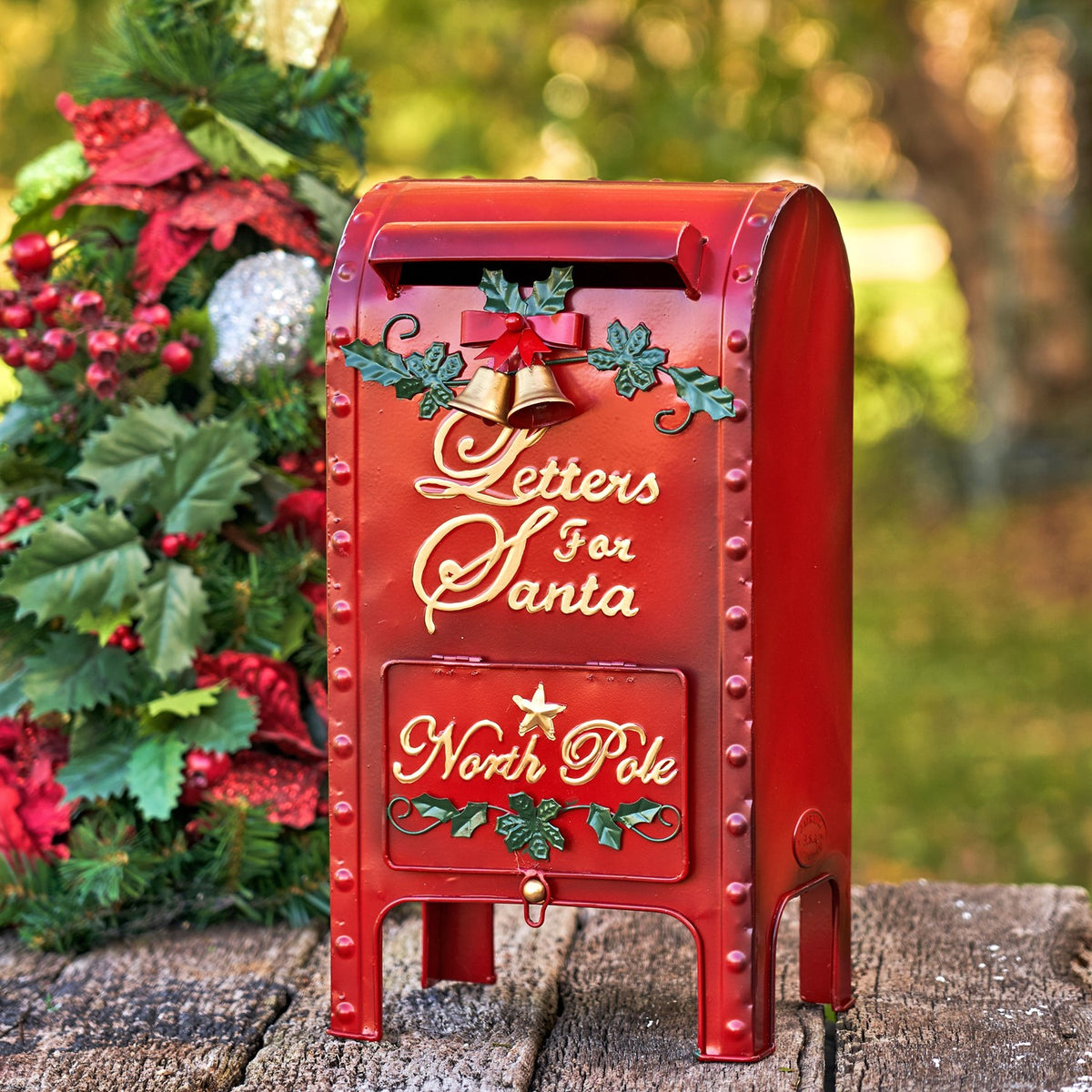 Red Christmas mailbox with a decorative bell and holly, standing beside a decorated tree.