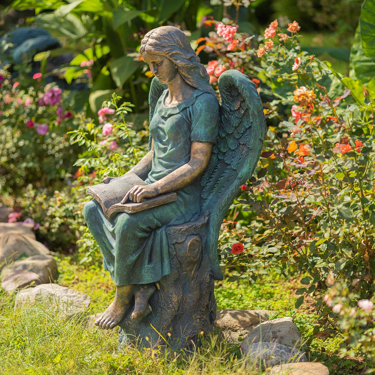 Teal-patina angel statue seated on a stump, reading an open book among garden flowers and greenery.