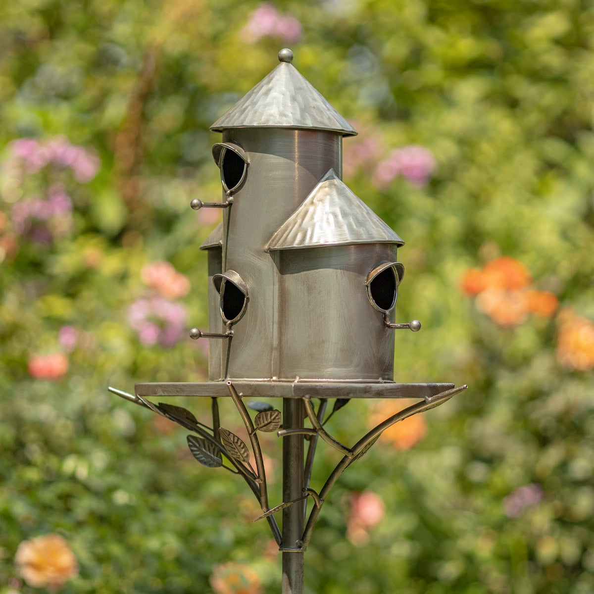 Silver triple birdhouse stake with three cylindrical houses, pointed roofs, and leaf-shaped metal supports in a garden.