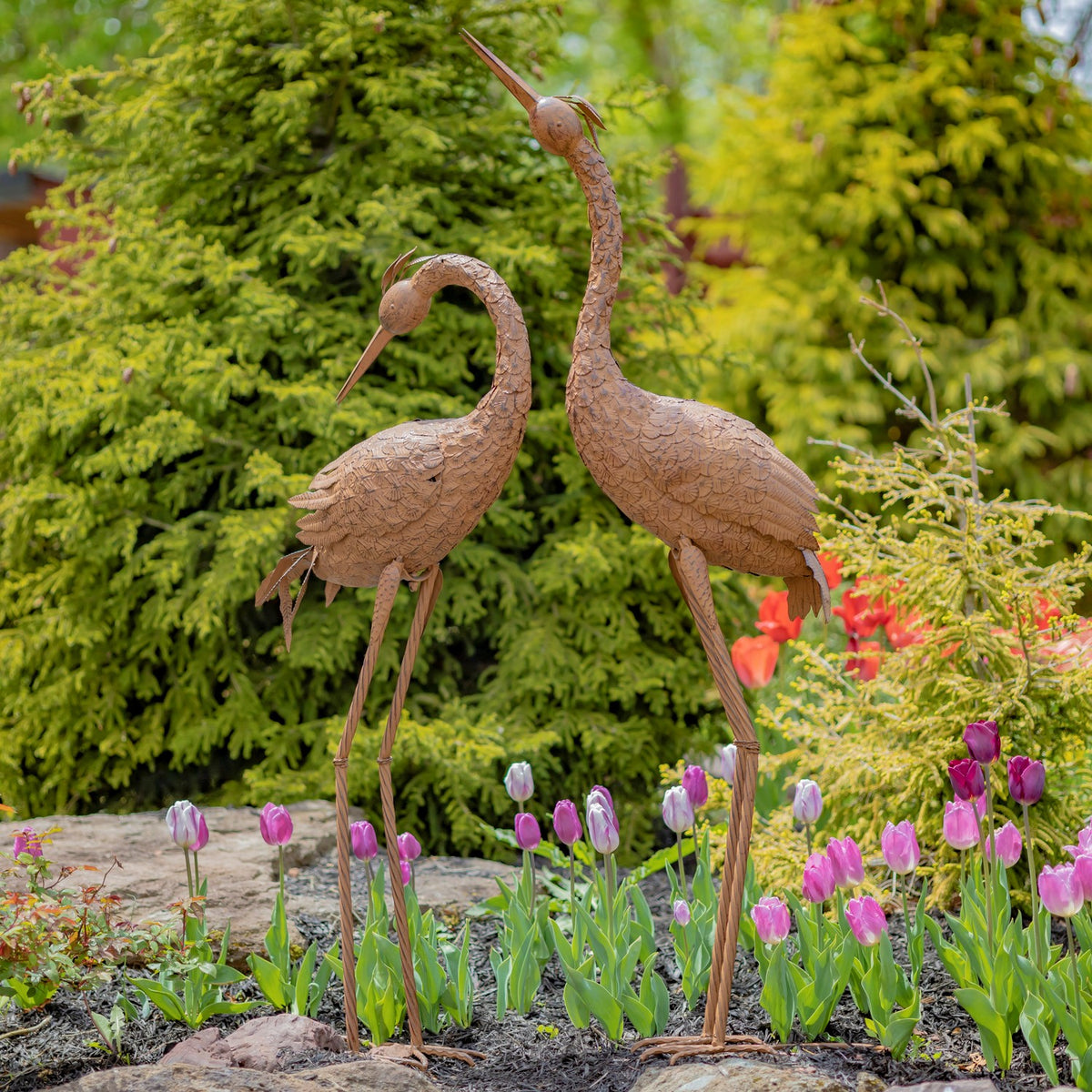 Pair of rust-brown crane sculptures standing among tulips in a garden