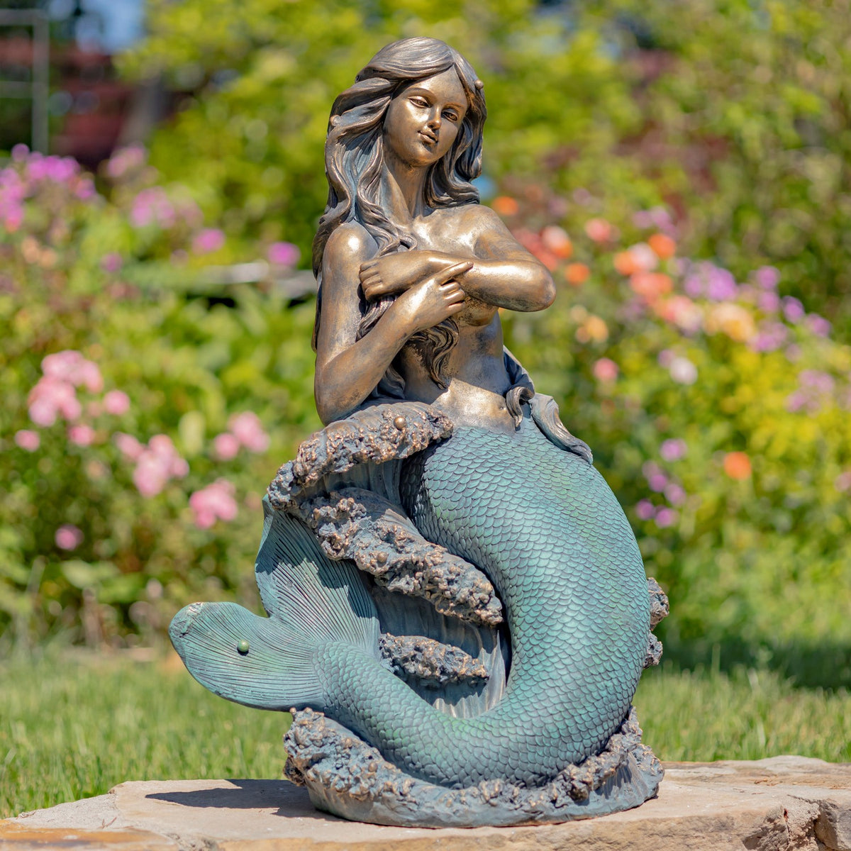 Mermaid statue seated on a rocky base, with a curled turquoise tail, displayed outdoors among flowers.