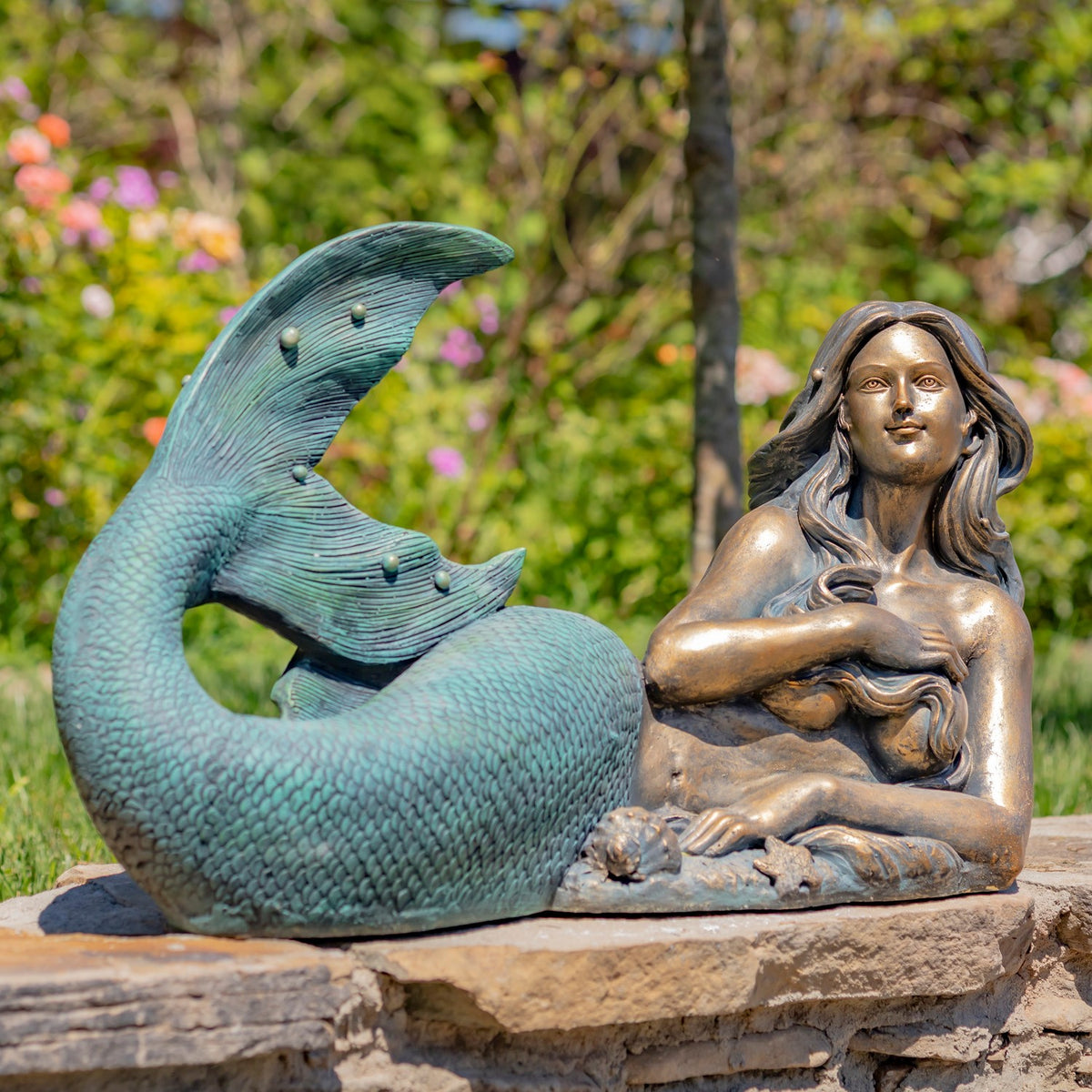 A reclining mermaid statue with a raised teal tail rests on a stone ledge in a garden.