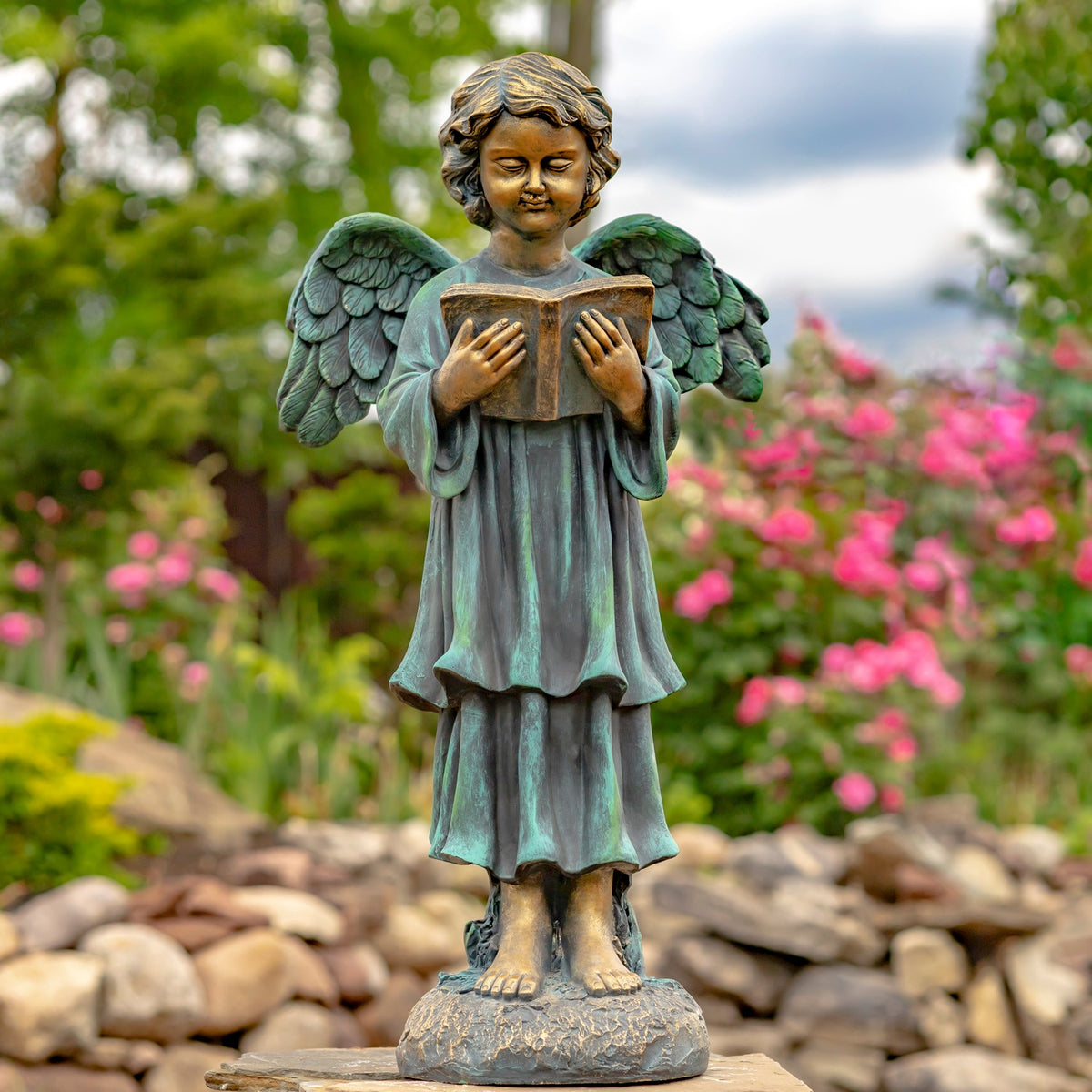 Dark green and bronze-toned angel statue reading an open book in a garden.