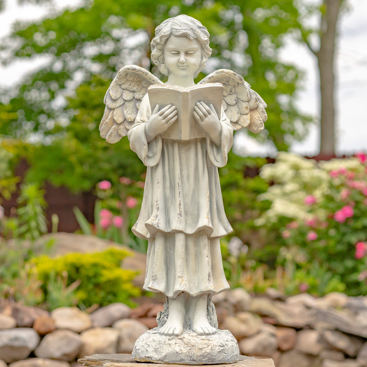 Light-colored angel statue standing among garden rocks, holding an open book beneath leafy trees.