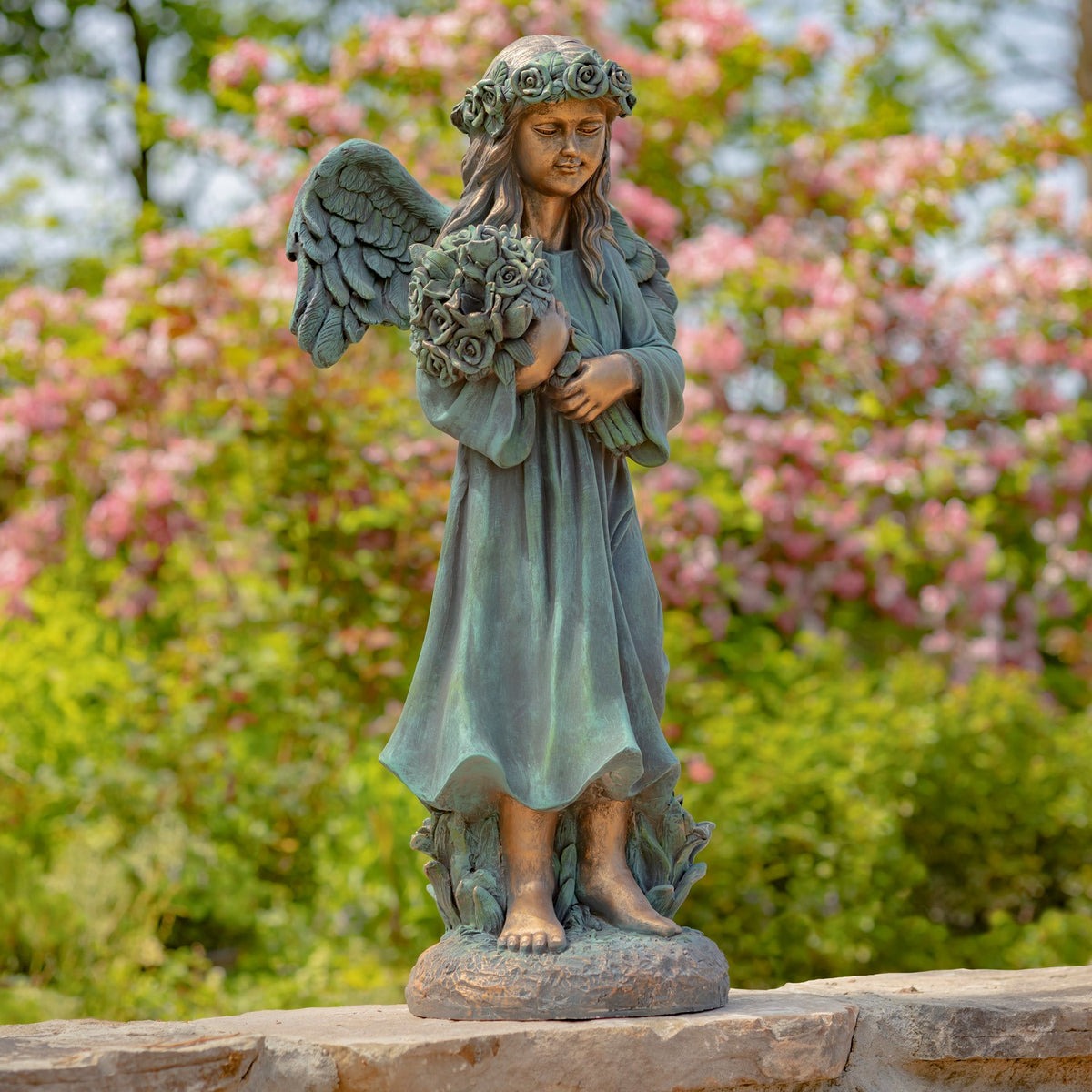 Winged statue in a long robe holds a bouquet of roses on a stone ledge amid flowering trees.