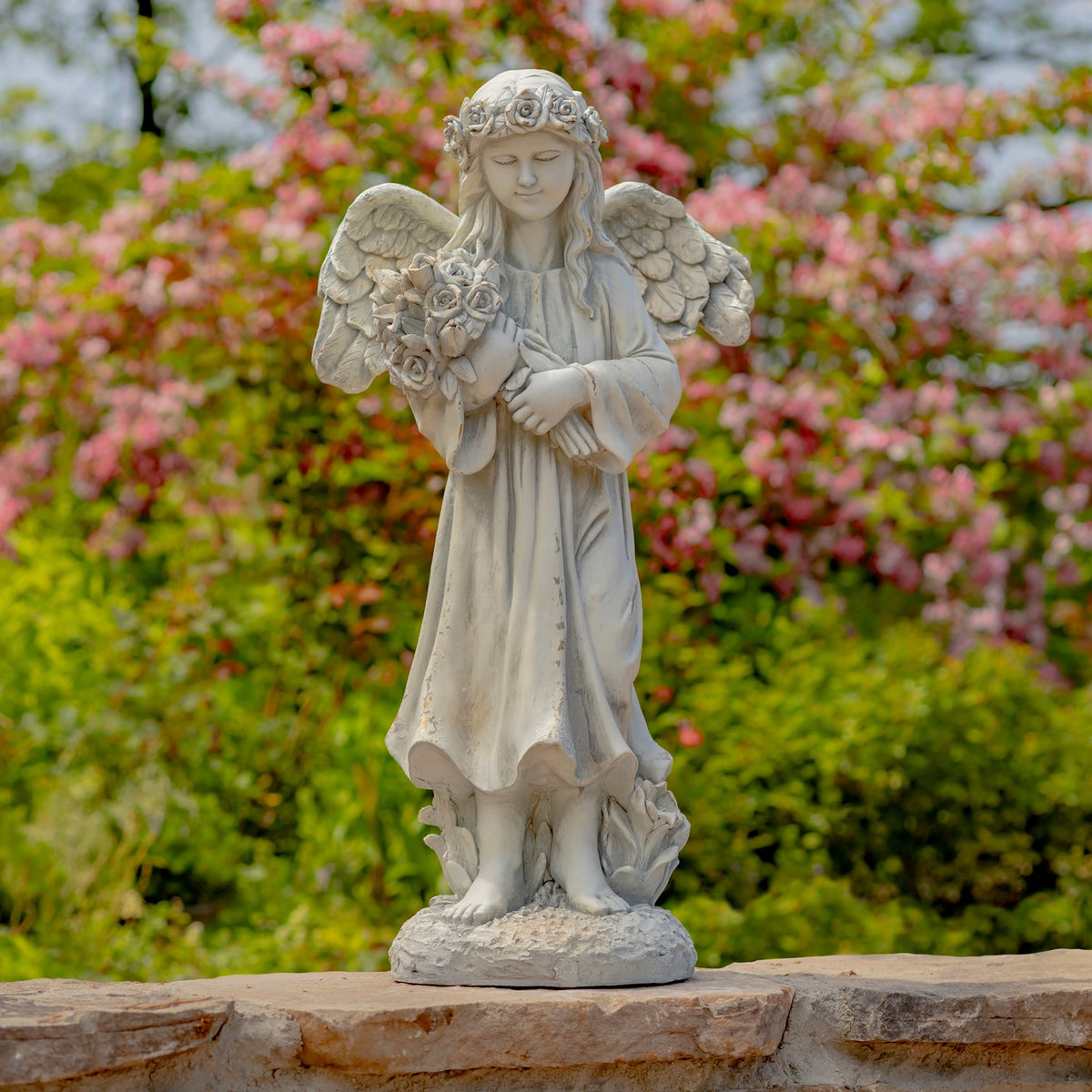A pale angel statue holding a bouquet stands on a stone wall amid flowering shrubs.