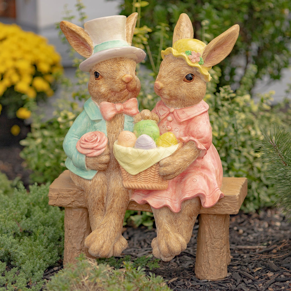 Two dressed rabbits sit on a rustic bench holding a basket of colorful eggs among garden plants.