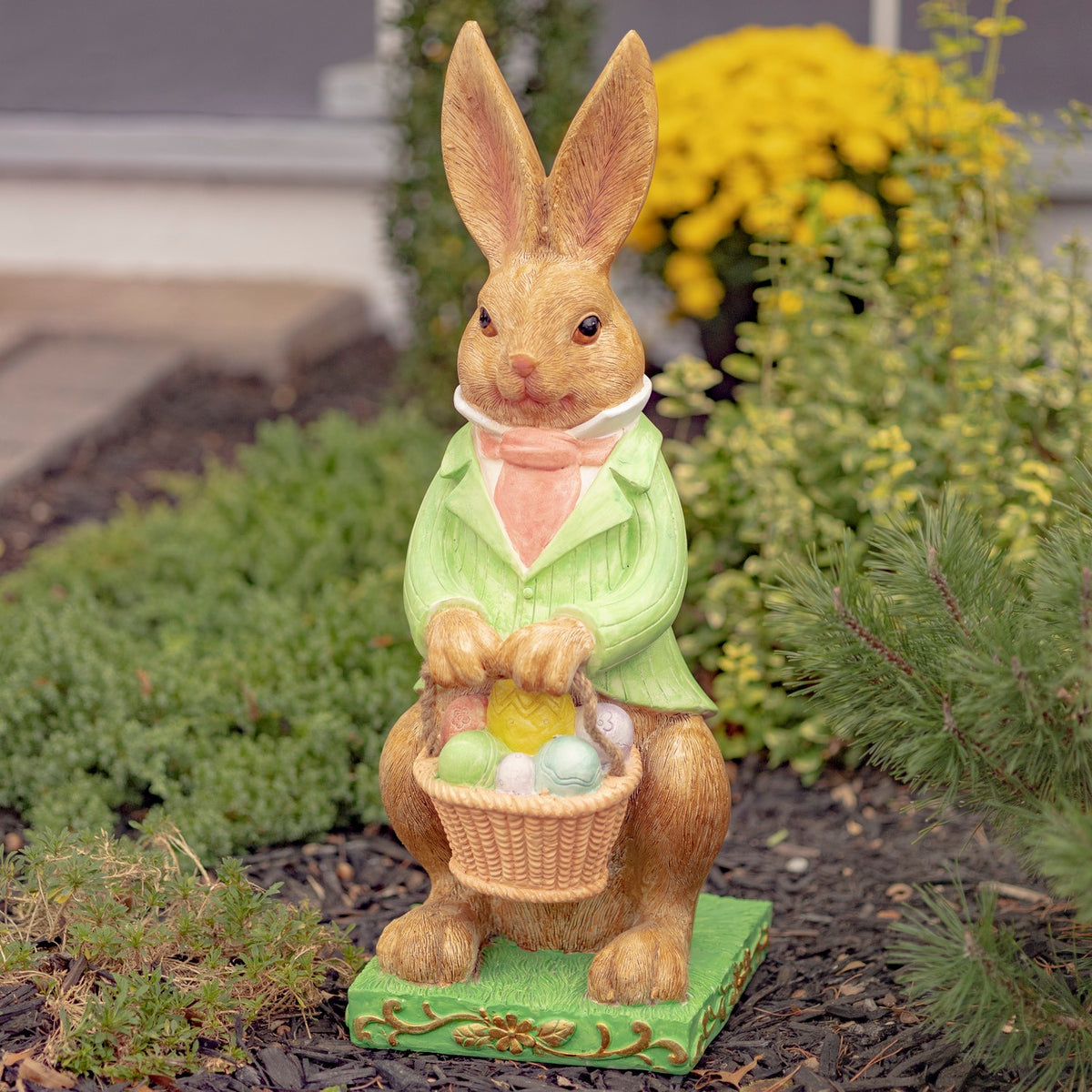 Rabbit statue in a green jacket holds a basket of pastel eggs among garden plants.