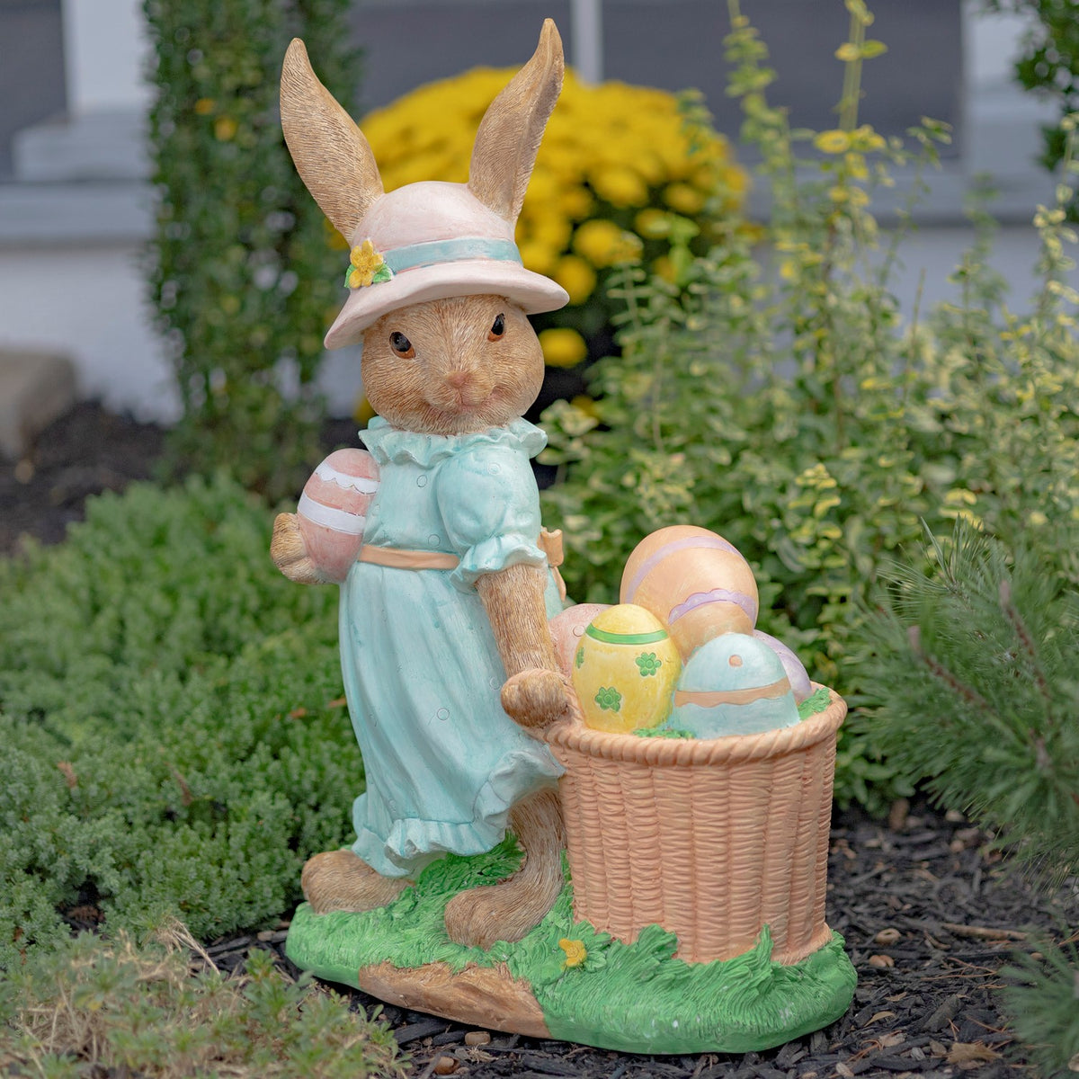 Rabbit statue in a pale blue dress and sun hat stands beside a basket of pastel eggs.