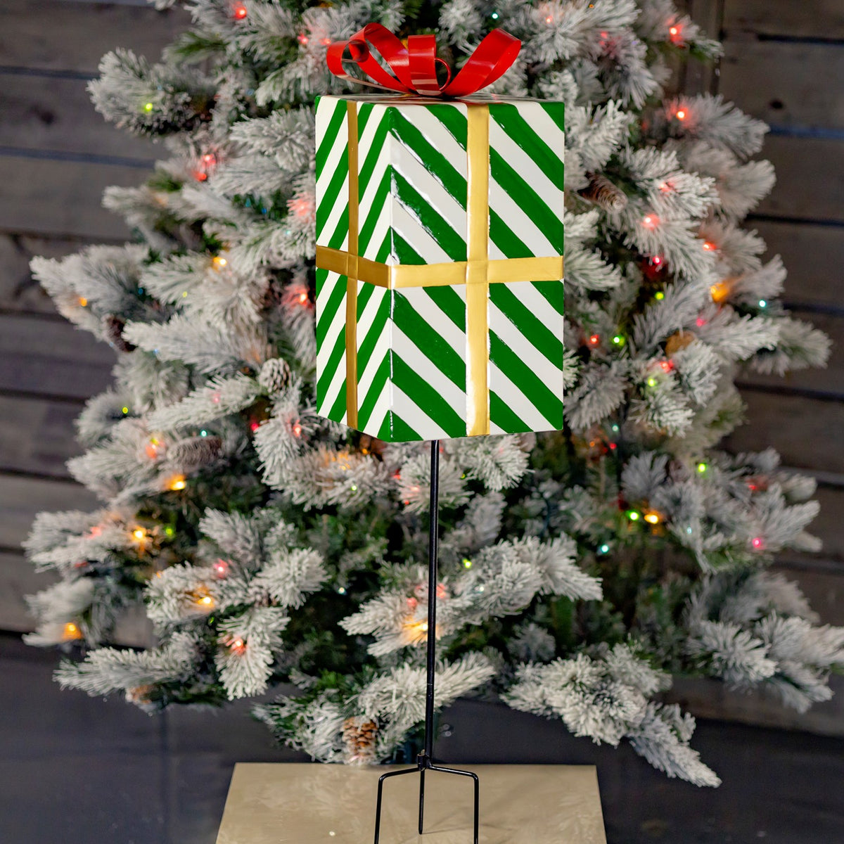 Green-and-white striped gift box stake with a red bow stands before a lit Christmas tree.