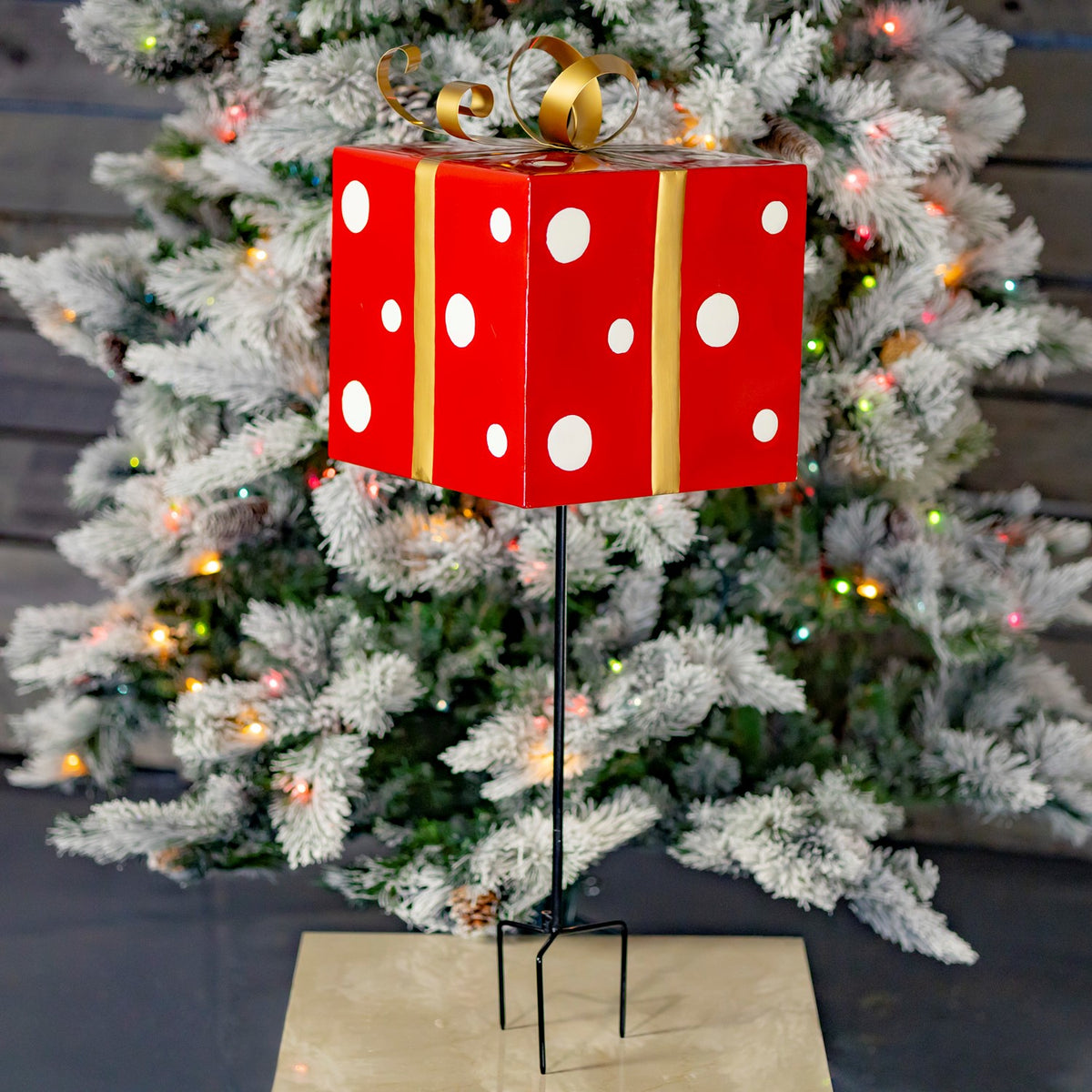 Red gift-box Christmas stake with white dots and a gold bow standing before a lit tree.
