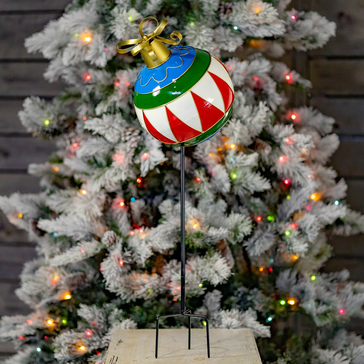 A red, white, green, and blue ornament stake stands before a lit Christmas tree.