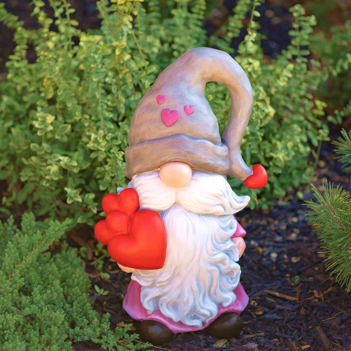 A gnome with a heart-decorated gray hat holds a large red heart among garden plants.