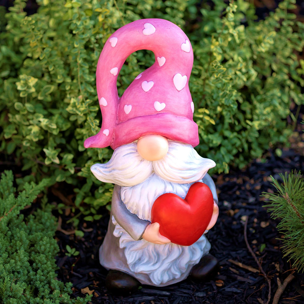 White-bearded gnome holding a red heart, front view among green garden plants